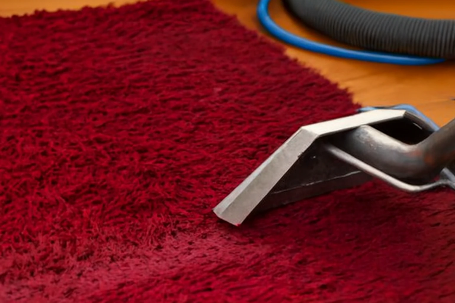 Steam cleaning process on a deep red shaggy carpet with visible areas of dirt. A silver cleaning wand emits steam as it moves across the rug, with a modern sofa in the background